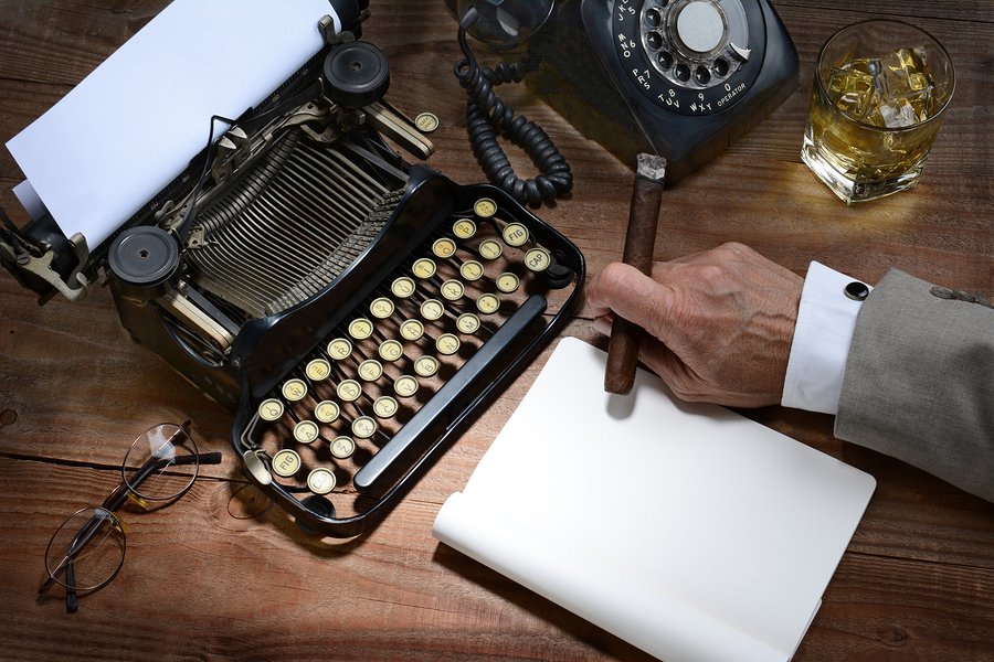 Closeup of a writer at his desk with a typewriter, rotary telephone, glass of whiskey and a cigar. A vintage feel with only the mans hand holding a cigar being shown.